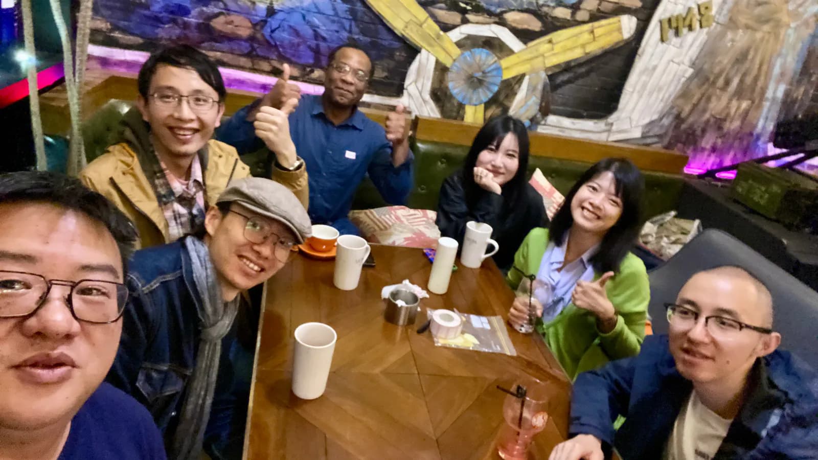 A selfie of seven of us around a cafe table covered in mugs, under a painted mural