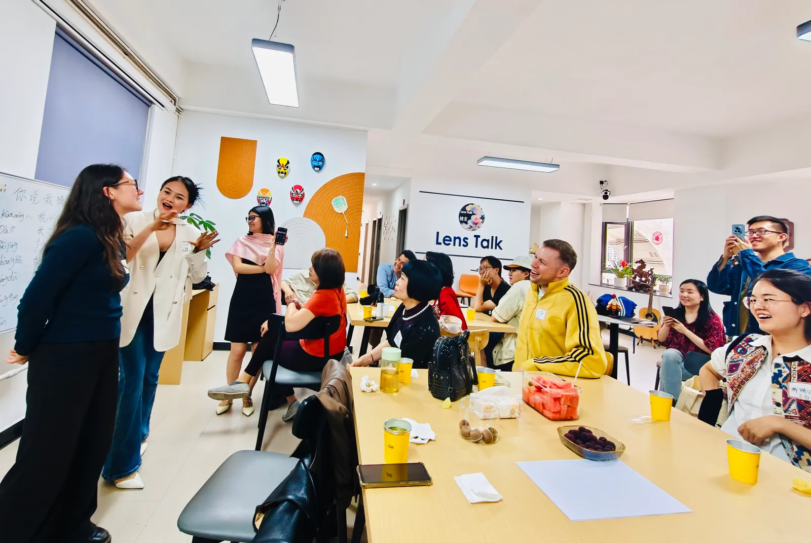 A guessing game at the whiteboard, city names in pinyin behind, the table laughing along