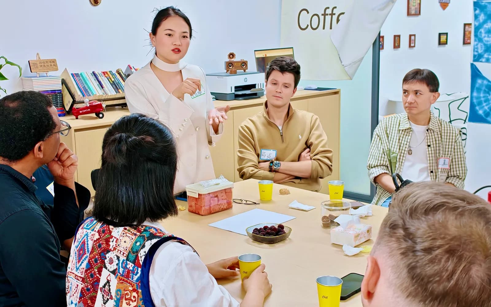 Someone standing to explain the round while the table listens over fruit and tea